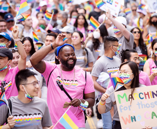 A diverse group of people marching in the San Francisco Pride Parade. They are wearing pink and grey t-shirts, waving their arms in celebration, and waving pride flags.