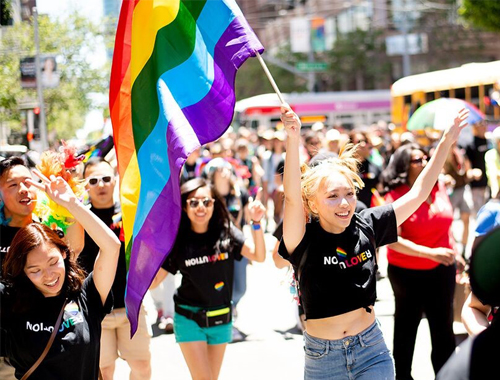 A group of people at the San Francisco Pride Parade looking very festive. The photo is centered on one person with blonde hair and a black t-shirt, who holding a large rainbow flag and is marching with joy.