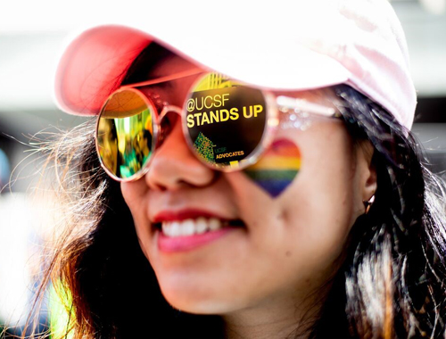 A person's face, close up. They have brown hair, are smiling and wearing a pink hat and sunglasses, and have a rainbow heart on their cheek. There is a reflection of a sign on their sunglasses that says, UCSF stands up.