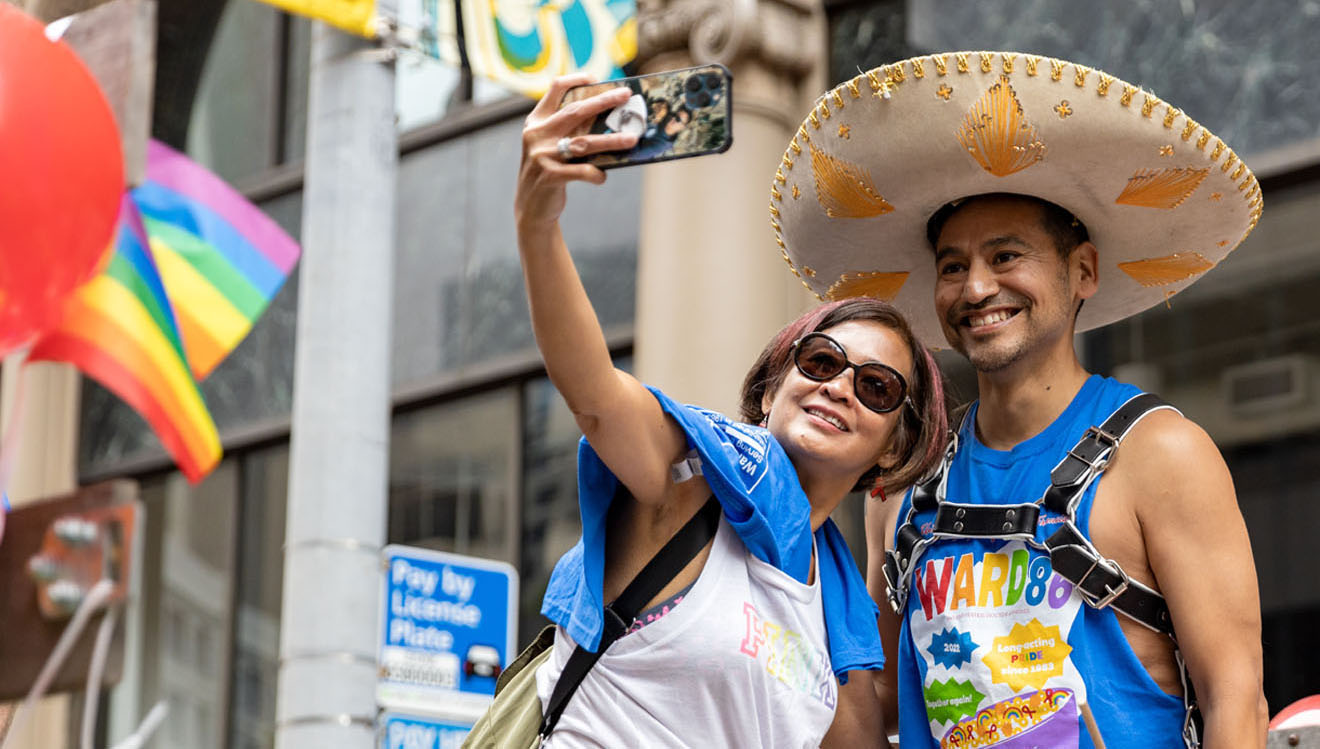A photo of two people of color taking a selfie together. One person is wearing sunglasses and holding their camera to take the photo. The other person posing is wearing a Mexican sombrero, a Ward 86 pride t-shirt and a black chest harness.